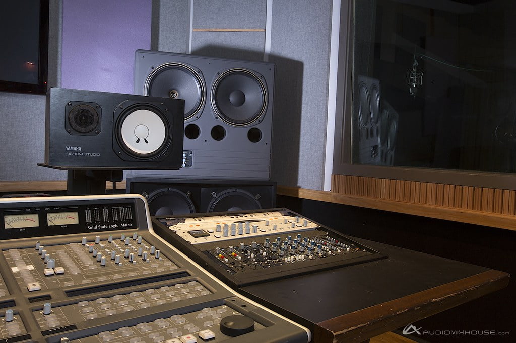 A professional audio mixing console and monitor speakers inside a recording studio, with a reflective window showing the silhouette of a microphone on a stand.