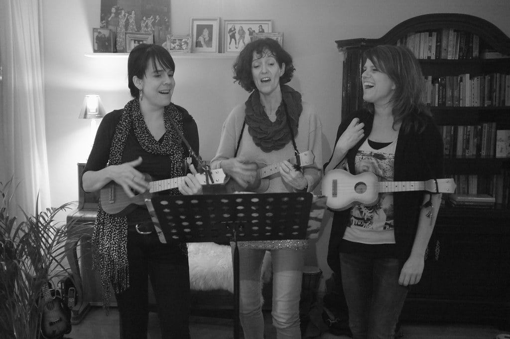 Three women singing and playing ukuleles in a living room with a bookshelf in the background; the image is in black and white.