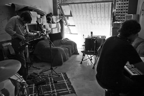 Two musicians practicing in a home studio with a guitarist standing and a keyboard player sitting, amidst instruments including a drum set, in a room with a patterned rug and a bed. The image is in black and white.