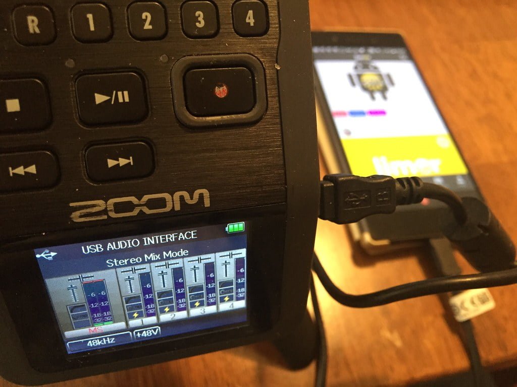 A close-up of a Zoom USB audio interface with various control buttons and a display screen showing "Stereo Mix Mode." A USB cable is plugged into the interface and leads to a smartphone with a blurred screen in the background.