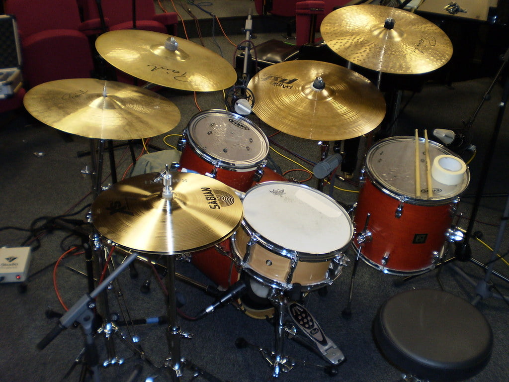 A red five-piece drum set with various cymbals, drumsticks on the snare drum, and a pedal, set up on a carpeted floor in a room with chairs in the background.