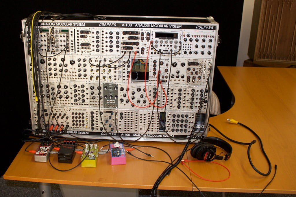 A Doepfer A-100 analog modular synthesizer with a complex array of patches and cables on a table, accompanied by various effect pedals and a pair of headphones.