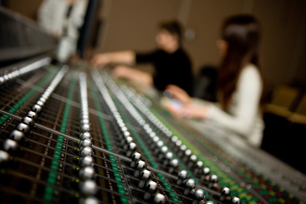 A close-up of an audio mixing console with numerous knobs and sliders in focus, while three people are blurred in the background, indicating they are working with the audio equipment.