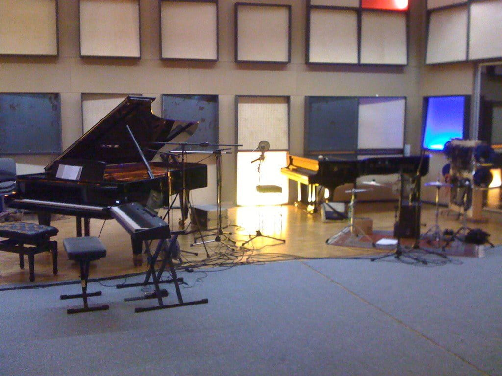 A music studio with two grand pianos, an electric keyboard, microphones, and various audio equipment cables on the floor.