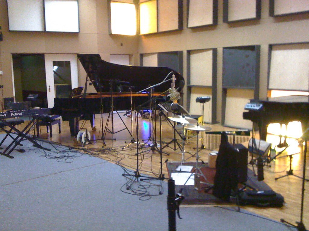 A music recording studio with a grand piano at the center surrounded by various microphones, music stands, an electronic keyboard, and assorted equipment.