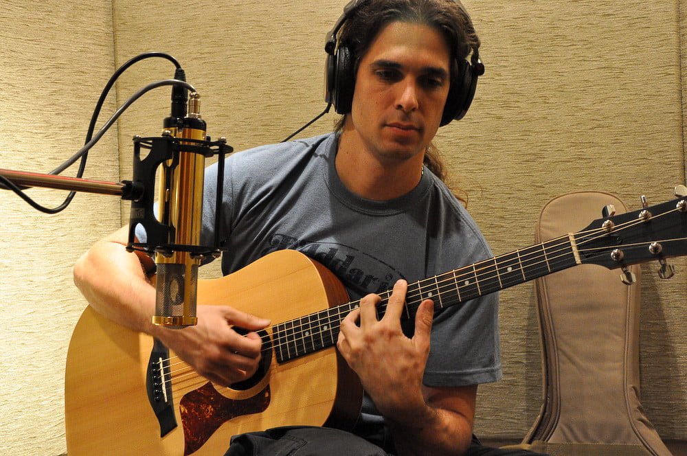 A person with headphones playing an acoustic guitar in a studio, with a large microphone positioned in front of them.
