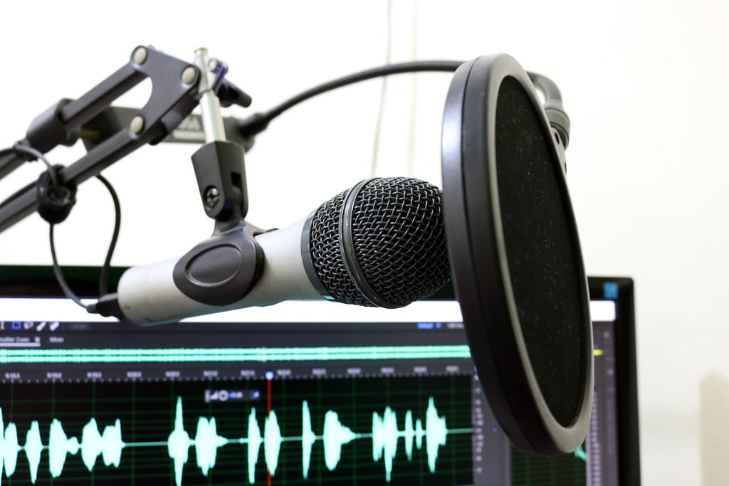 A professional microphone with a pop filter attached to a swivel arm stand, in front of a computer screen displaying audio waveforms.