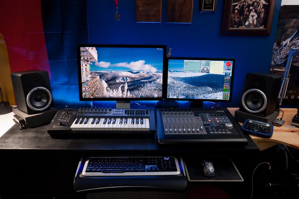 A home music production setup with two monitors showing desktop and flight simulator, studio monitors, a MIDI keyboard, an audio mixer, and a computer keyboard and mouse on a desk.