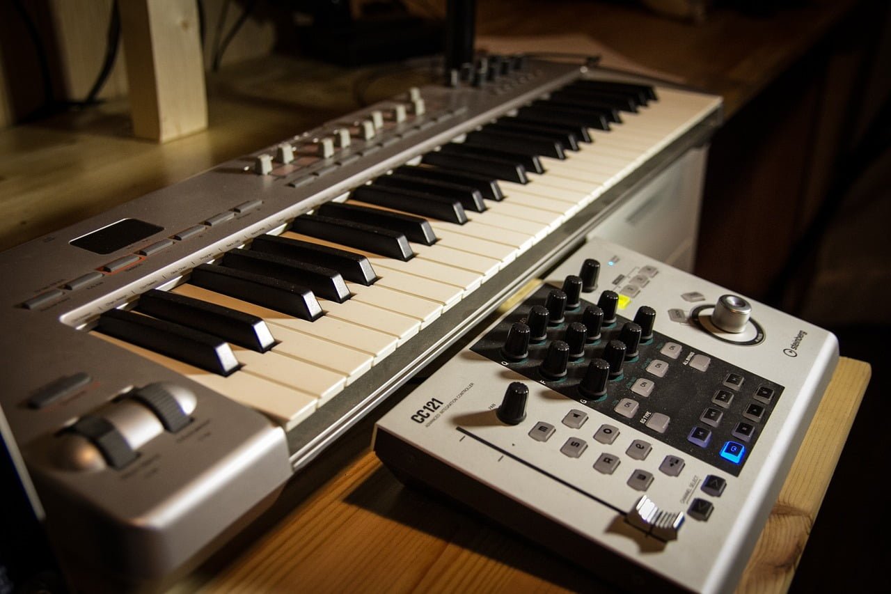 A MIDI keyboard controller with black and white keys next to a drum machine with knobs and pads on a wooden surface.