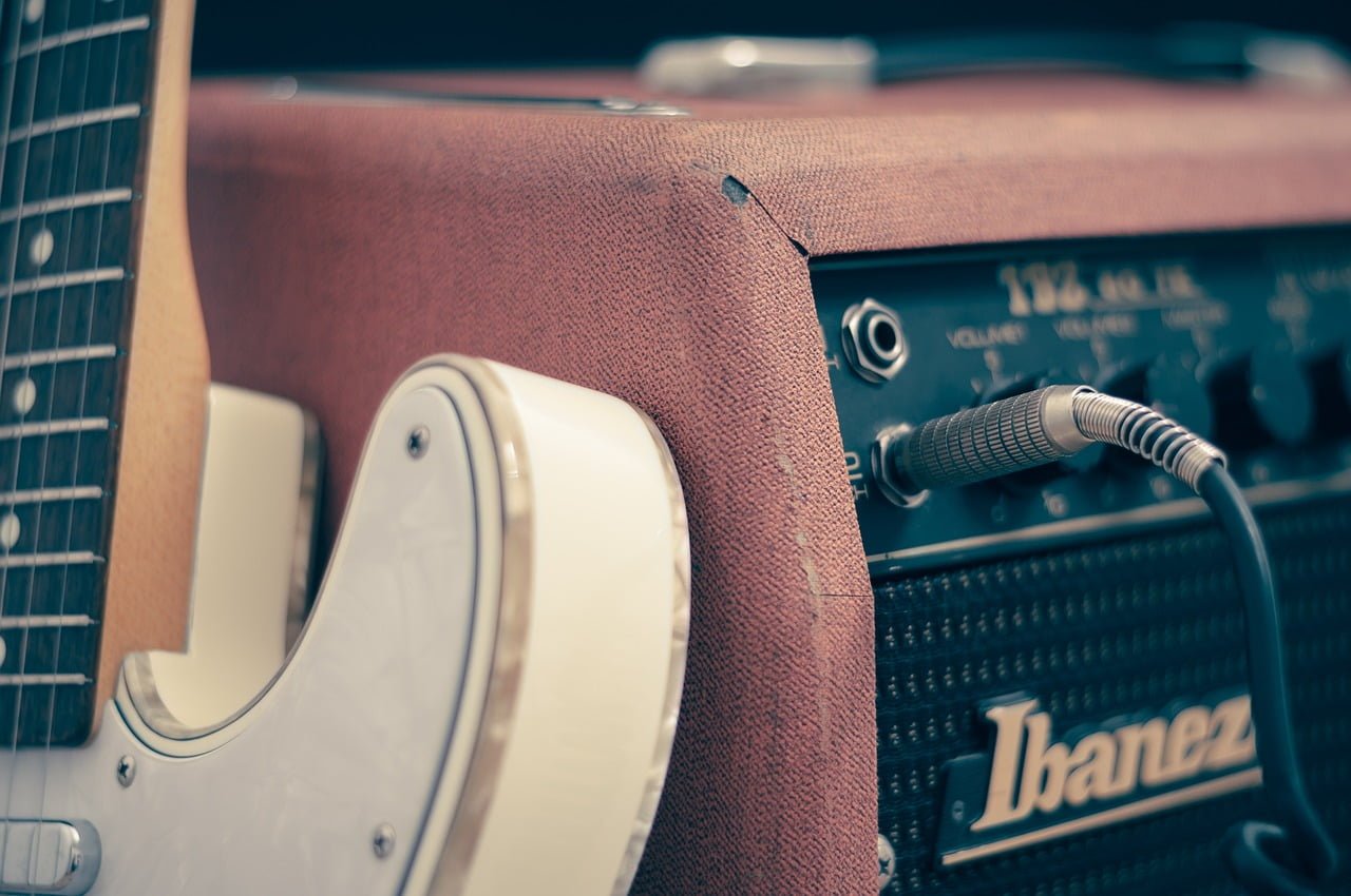 Close-up of a white electric guitar leaning against a brown Ibanez guitar amplifier with a cable plugged in.