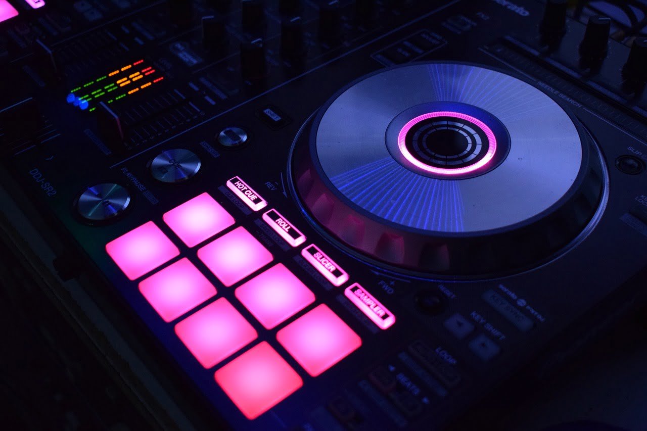 A close-up of a DJ mixing console with illuminated pads and jog wheel in a dark setting, featuring neon pink and blue lighting.