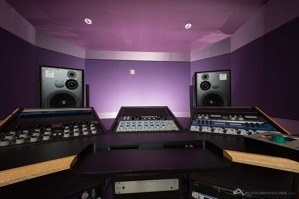 A professional music studio control room with sound mixing desks, rack-mounted audio equipment, and large monitor speakers in a room with purple acoustic treatment on the walls.