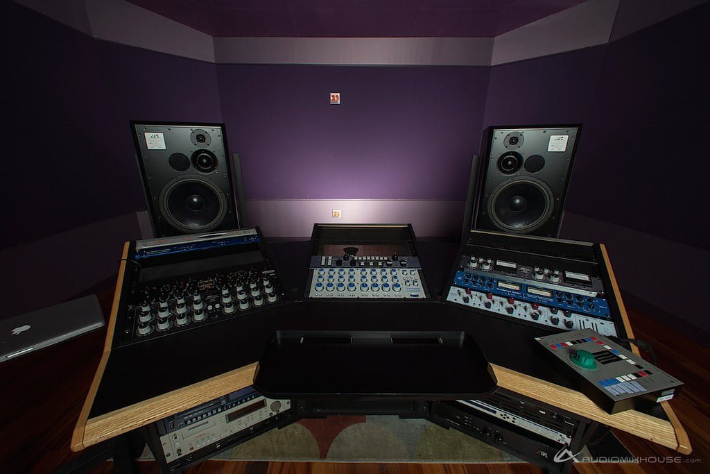 A professional audio mixing and mastering studio setup with large monitor speakers on either side of a mixing console, outboard gear racks, and a computer on a wooden desk in a room with purple walls.