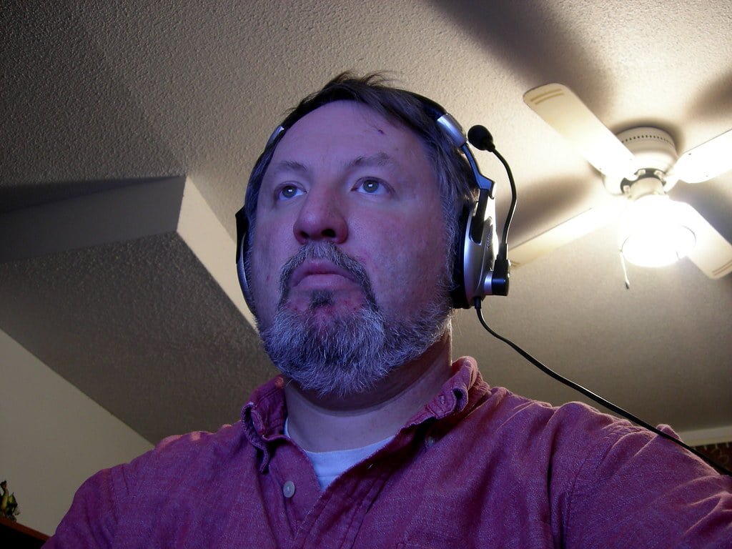 A man with a beard looking off to the side, wearing headphones and a red shirt, with a ceiling fan and light in the background.