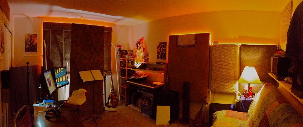 A panoramic view of a cozy, dimly lit home music studio with recording equipment, a computer, musical instruments, soundproofing panels, and decorative items.