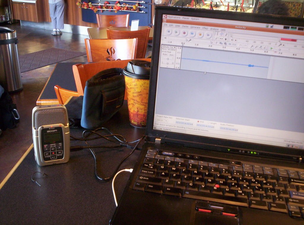 A laptop displaying an audio editing program on its screen, with a portable audio recorder and a cup of coffee on a cafe table.