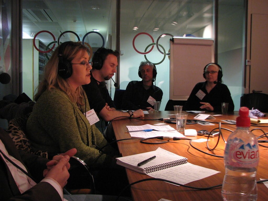 A group of people wearing headphones sitting around a conference table in a meeting room, with microphones and notepads in front of them, and an Evian water bottle in the foreground.