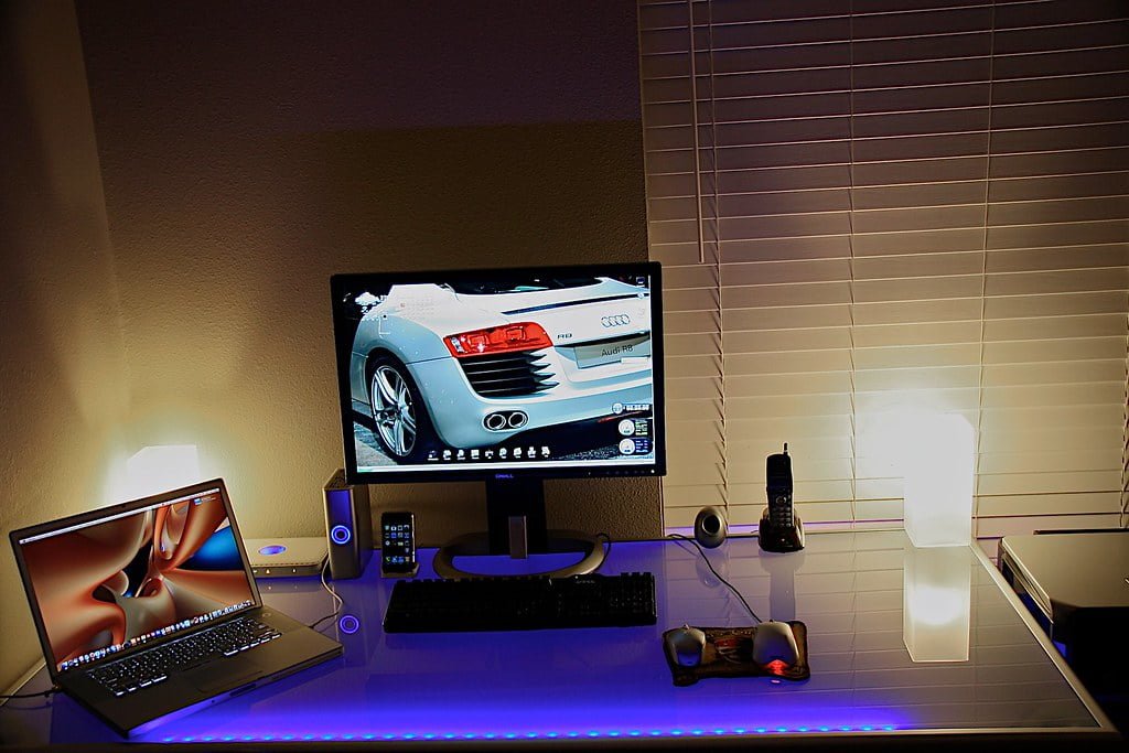 A modern home office setup with ambient lighting, featuring a laptop on the left, a desktop monitor with a car wallpaper, external speakers, a desk phone, a table lamp, and various accessories on a tidy desk.