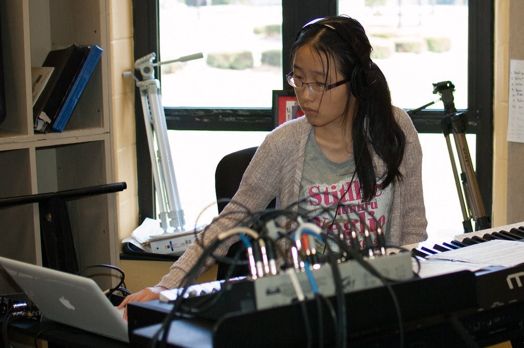 A young woman wearing headphones focused on using a laptop with musical equipment and a keyboard around her in a studio setting.