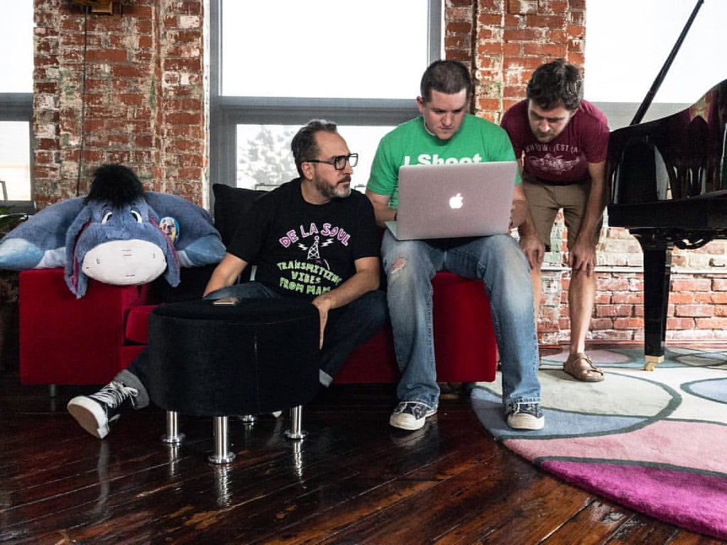 Three men in a casual setting with brick walls and a grand piano, one seated on a red chair looking at a laptop screen, the second seated beside him wearing a green shirt, and the third standing to the right, peering over the laptop screen. A large stuffed Eeyore toy is resting on the chair to the left.