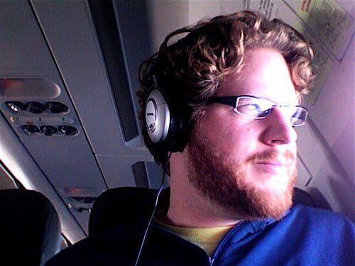 A man wearing headphones looks out the window on an airplane, with the interior of the cabin visible in the background.