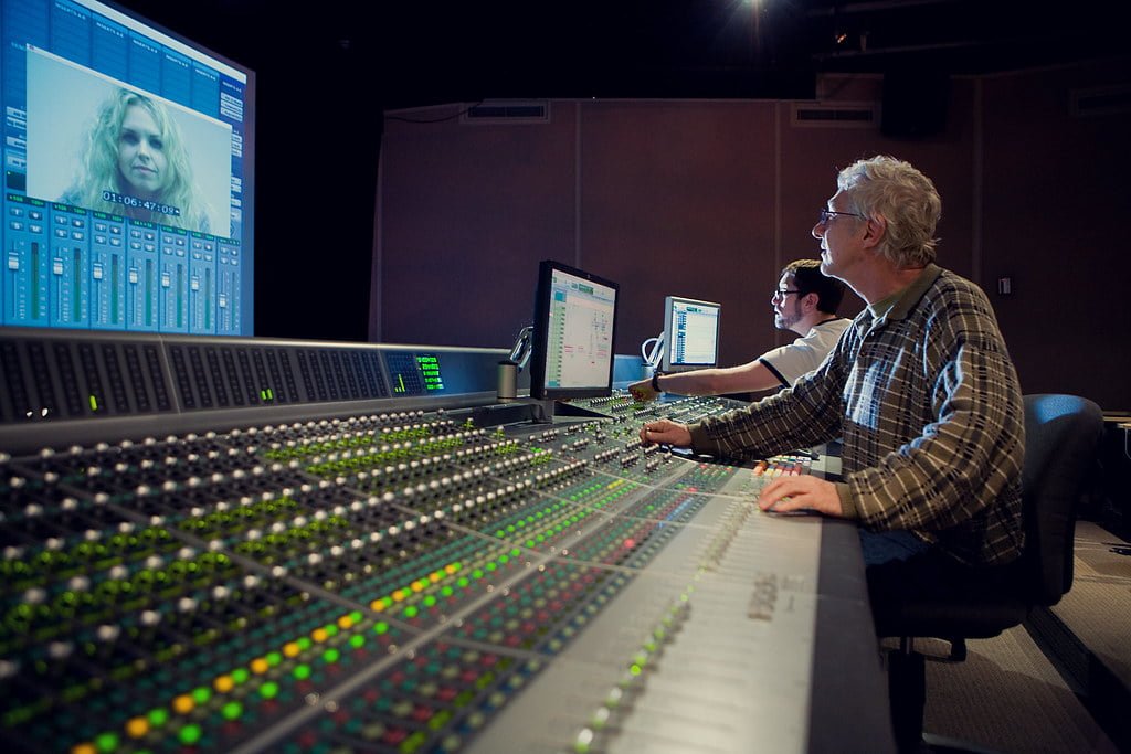 Two men operating a large audio mixing console in a studio with multiple computer screens displaying audio editing software and a video of a woman's face.