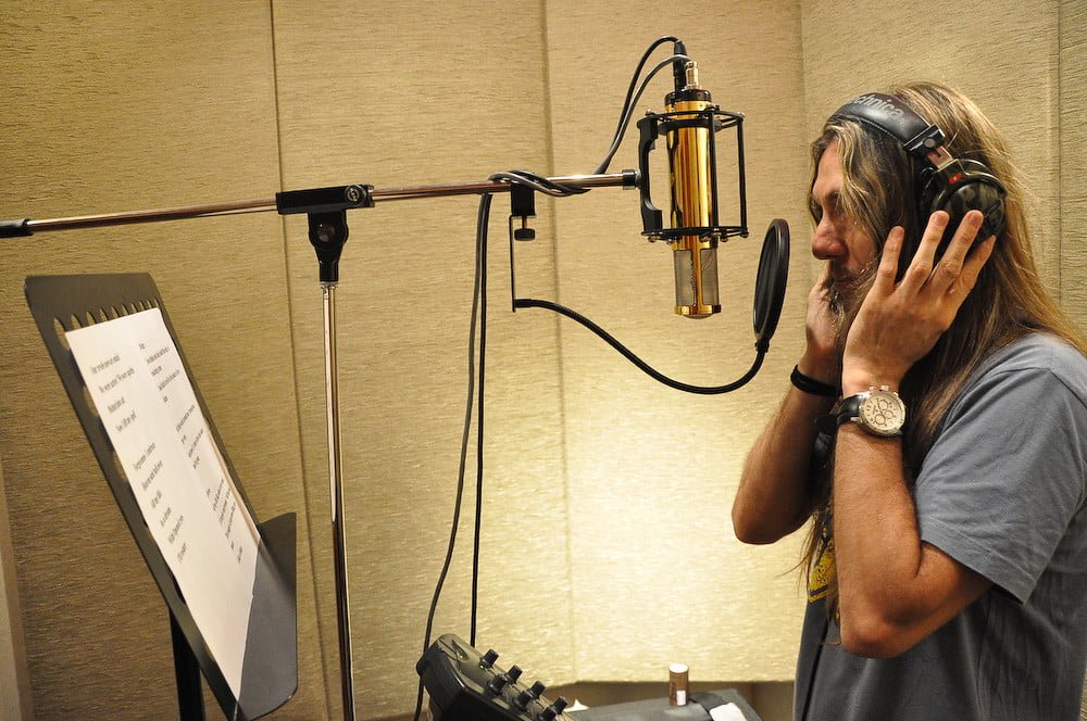 A person with long hair wearing headphones is standing in a recording studio, holding headphones with one hand, and looking at a microphone and a pop filter, with a sheet of paper with lyrics on a music stand.