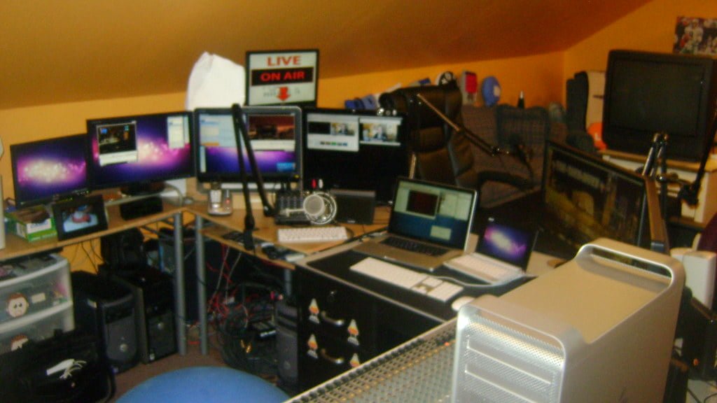 A cluttered home office with multiple computer monitors, laptops, microphones, and studio equipment, and a "LIVE ON AIR" sign illuminated in the background.