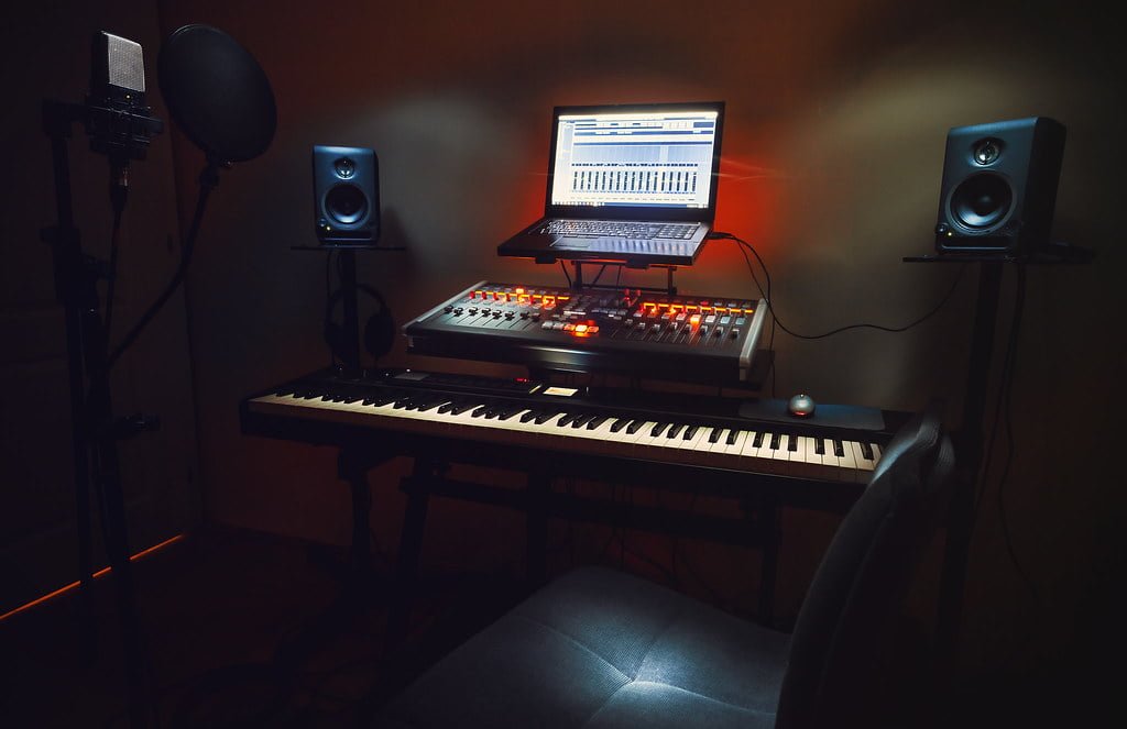 A home music studio setup with a keyboard, audio interface, studio monitors, a microphone with a pop filter, headphones, and a computer with digital audio workstation software on the screen, all in a dimly lit room with ambient lighting.