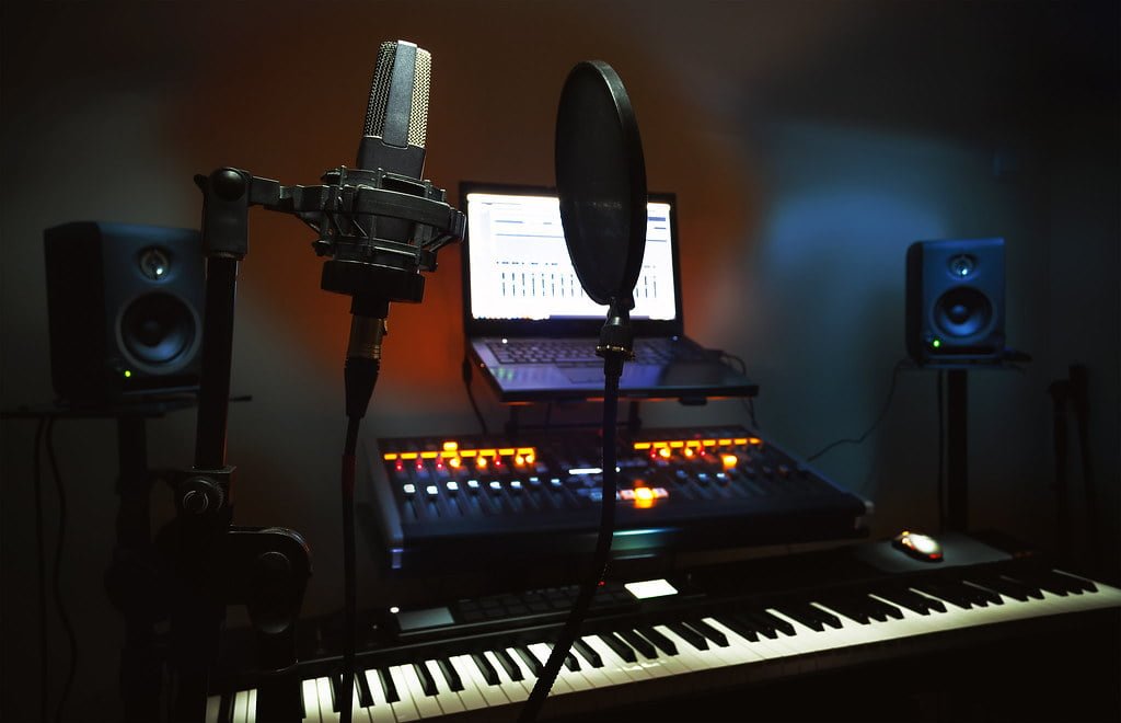 A home music studio setup with a condenser microphone in the foreground, a laptop displaying audio production software, an electronic MIDI keyboard, an audio mixing console with lit buttons, and two monitor speakers in a dimly lit room.