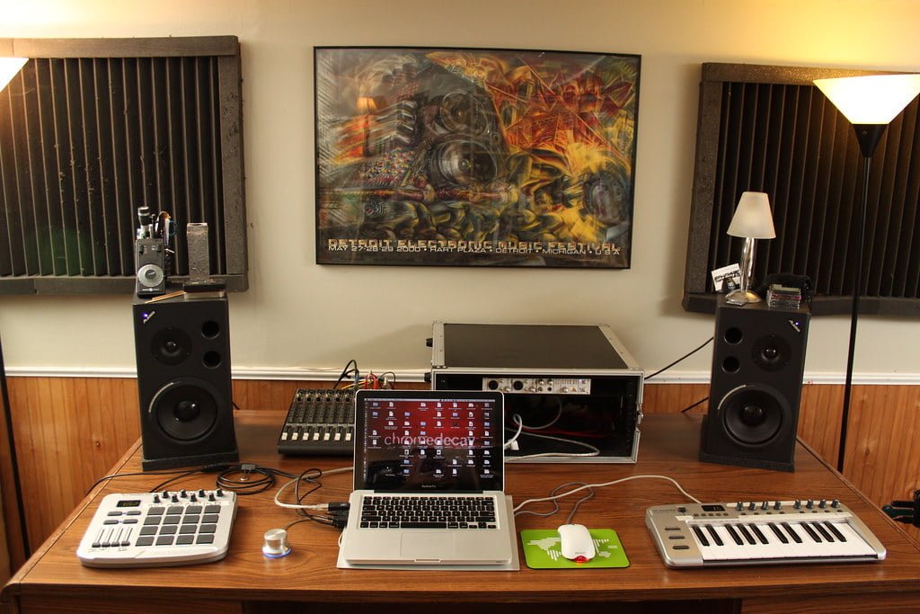 A home music studio setup with two large speakers flanking a wooden desk, which holds a laptop, MIDI keyboards, an audio mixer, and various recording equipment with soundproofing panels on the walls and a framed abstract painting above the desk.