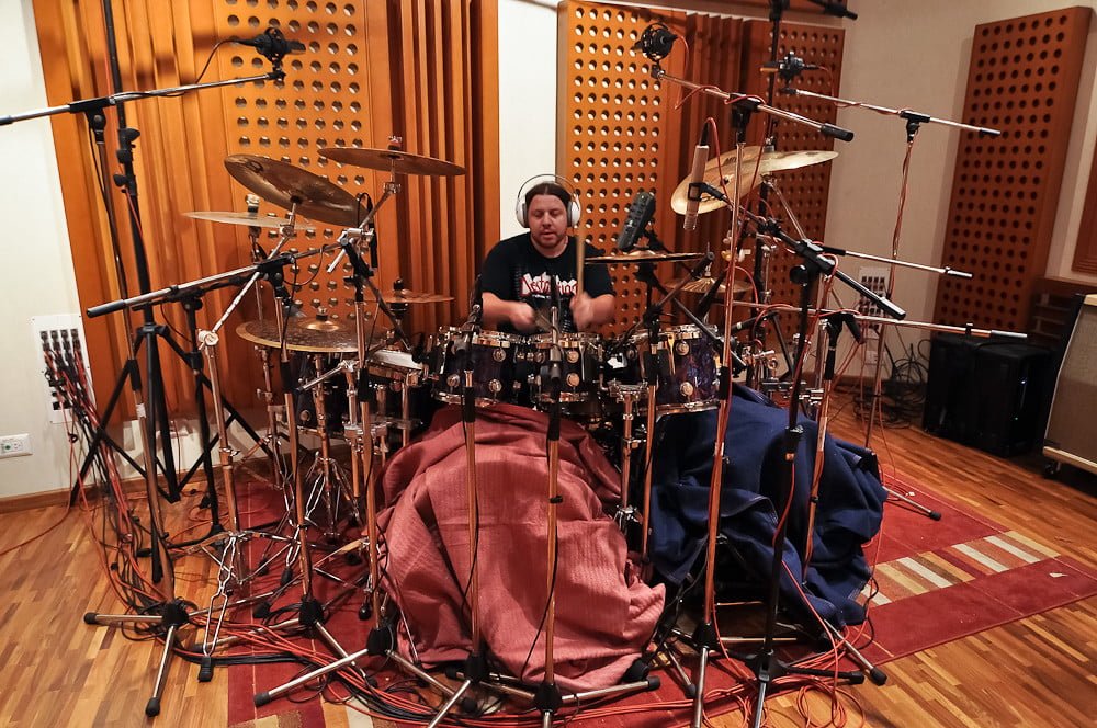 Drummer seated at an elaborate drum kit surrounded by microphones in a recording studio with acoustic paneling on the walls.