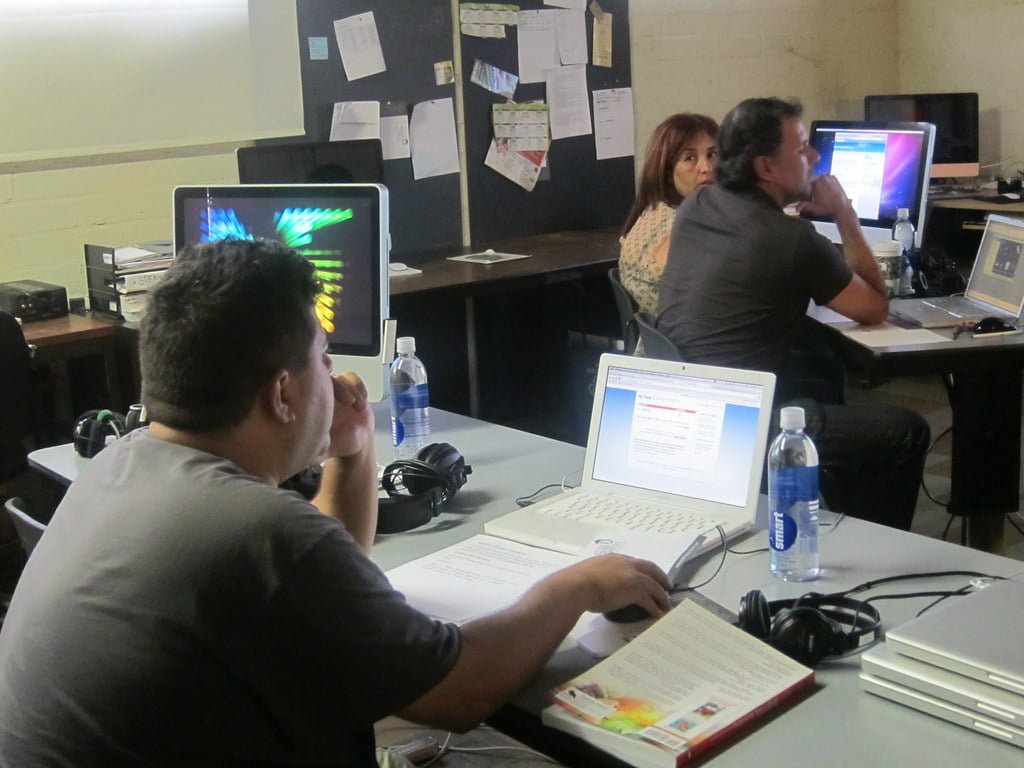 Adults in a computer class working on desktops and laptops in a casual, cluttered classroom setting.