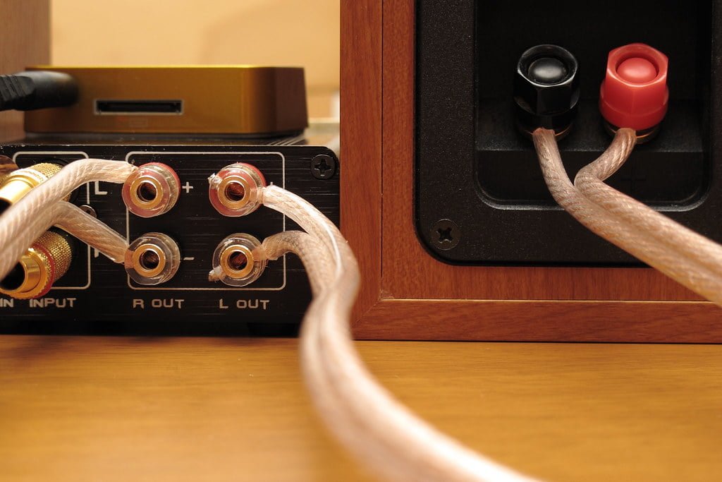 Close-up of high-fidelity audio equipment showing speaker terminals with transparent-insulated cables connected, and part of an amplifier on a wooden surface.