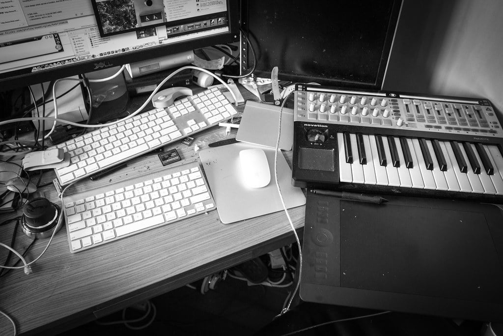 A black and white photo of a cluttered workspace with two computer keyboards, a small MIDI keyboard, headphones, a drawing tablet, and various cables.