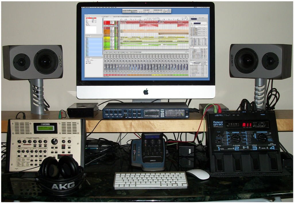 A home music studio setup with a computer displaying audio production software, flanked by two studio monitor speakers, an audio interface, various rack units, and a pair of headphones on a desk.