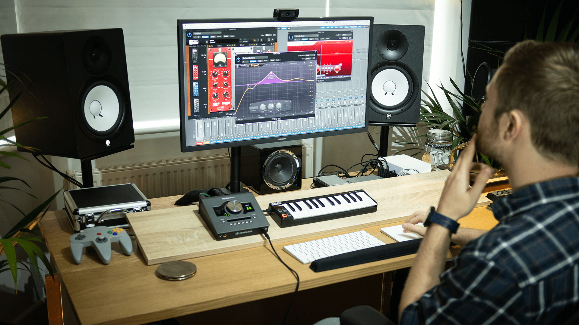 A person is working in a home music studio with professional audio equipment, including studio monitors, an audio interface, and a MIDI keyboard, while looking at a computer screen displaying music production software.