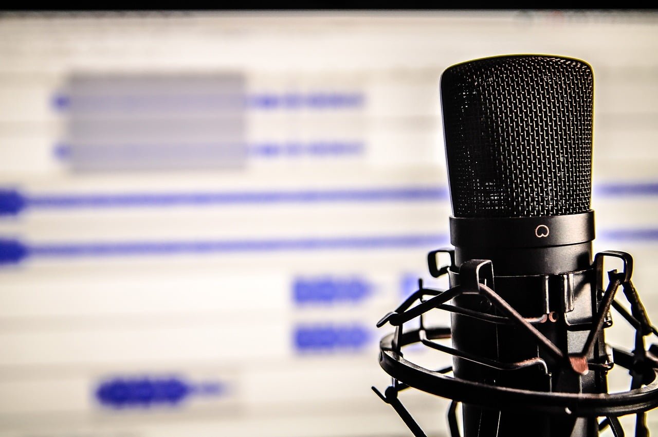 A close-up of a black microphone with a shock mount set against a blurred background of computer code on a screen.