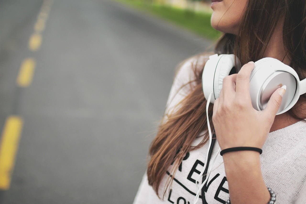 Woman holding white headphones on her shoulder by a roadside, with the focus on her and the headphones, and the background softly blurred.