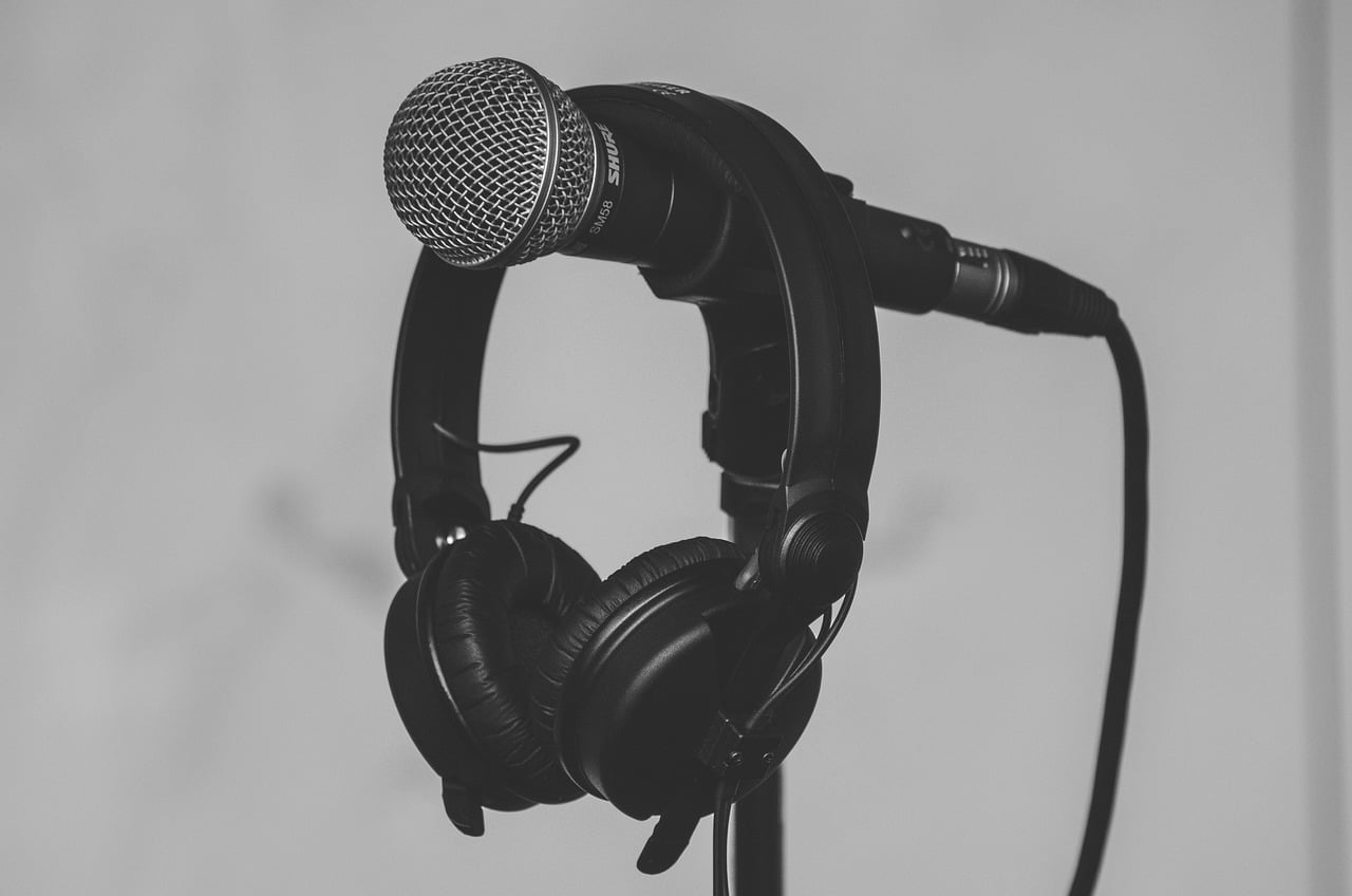 A black and white image of a microphone with a mesh head attached to a stand, with over-ear headphones hanging off the microphone stand.