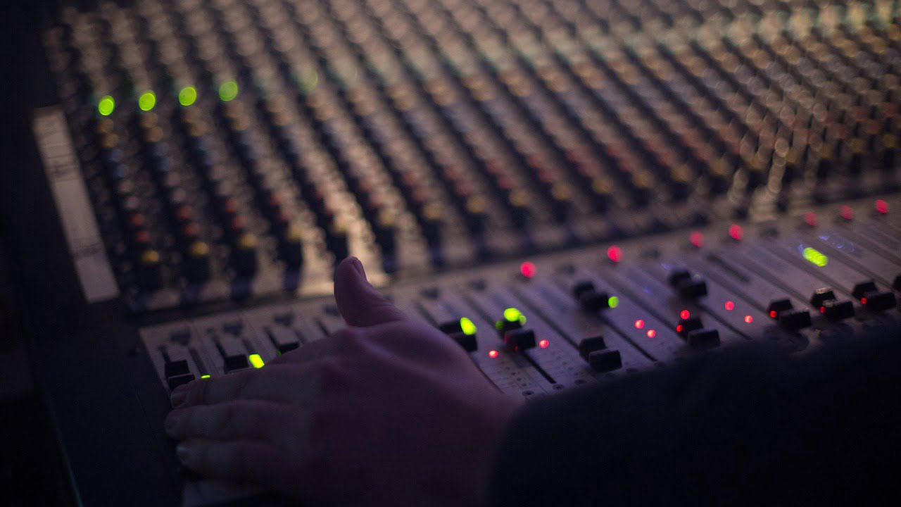 A person's hand adjusting sliders on a sound mixing console with illuminated buttons under low light conditions.