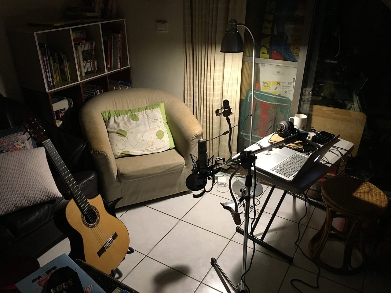 A cozy home recording studio setup at night with a laptop on a table, audio interface, microphone on a stand, headphones, a classical guitar leaning against a chair, and a bookshelf in the background, all illuminated by a floor lamp.