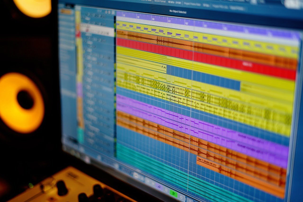 Close-up of a digital audio workstation screen showing colorful track layers with audio waveforms and editing tools, with a studio monitor speaker blurred in the background.