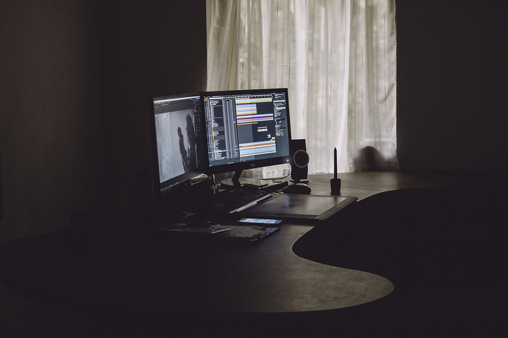 A dimly lit room with a dual-monitor computer setup on a desk, displaying graphics software. Light filters through sheer curtains behind the desk. There's a smartphone, a notebook, and a speaker on the desk as well.