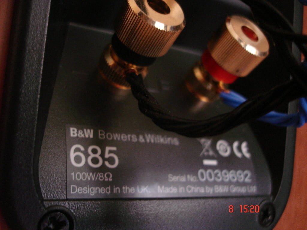 Close-up of the back panel of a Bowers & Wilkins 685 speaker, showing its connections and specification label indicating a power rating of 100W/8Ω. The image is slightly out of focus, with the timestamp '8 15:20'.