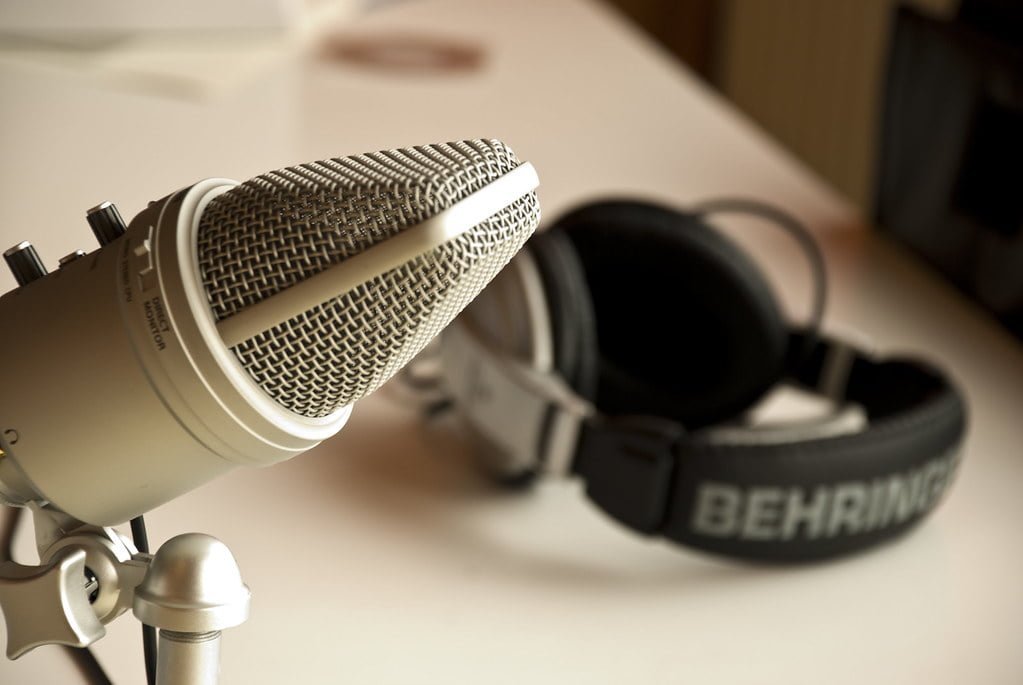 A close-up of a metallic studio microphone in focus, with a pair of headphones in the background displaying the brand 'BEHRINGER'.