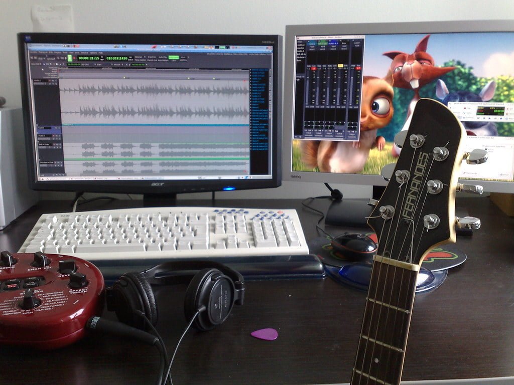 A home recording studio setup with audio editing software visible on the monitor, a red electric guitar leaning against the desk, and headphones lying on the guitar's body. Another monitor displays an animated squirrel character.