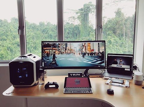 A tidy workspace with a large ultrawide monitor displaying a city street view, a smaller laptop screen, external speakers, a digital clock showing "4:44 PM," a smartphone, and a mouse, with a forest view outside the window.