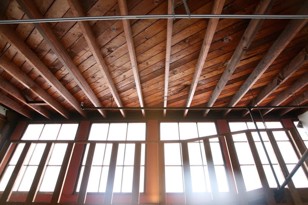 Interior view of a wooden ceiling with exposed beams above large windows with a metal grid design.