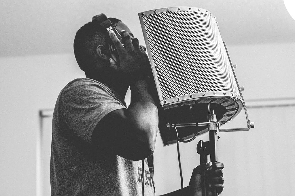 A black and white photo of a person standing before a studio microphone and pop filter, appearing to be singing or speaking with their hand on the headphones covering their ears.